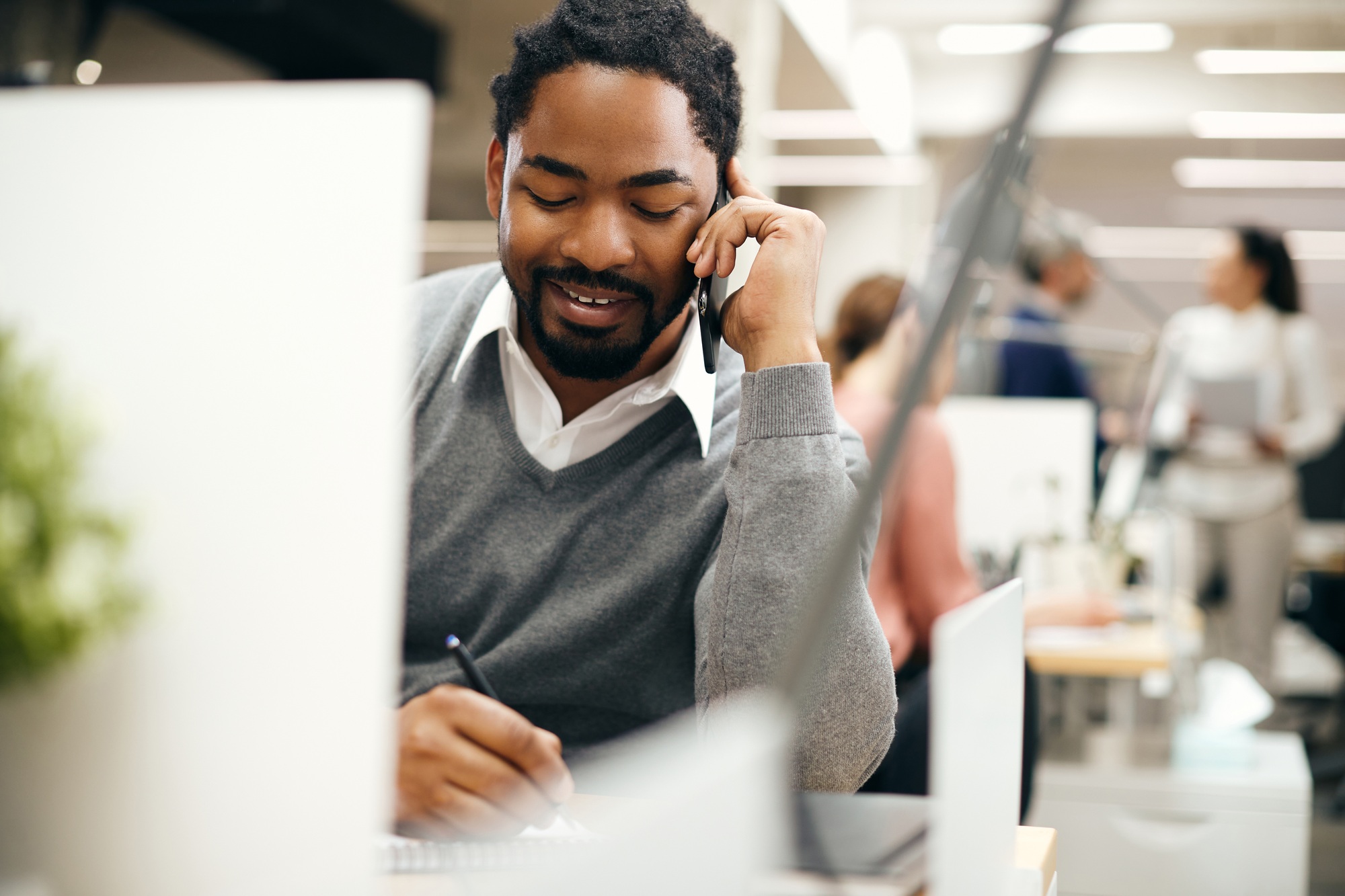 Happy black businessman talking on the phone and writing notes while working in the office.